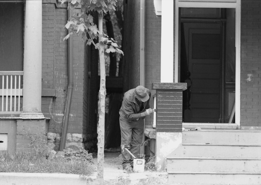 A person in a cap painting a brick porch pillar on a house