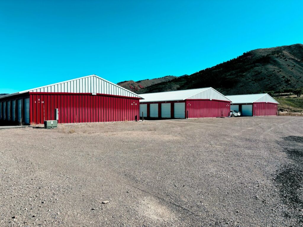 Three red and white storage unit buildings on a gravel lot before rolling hills